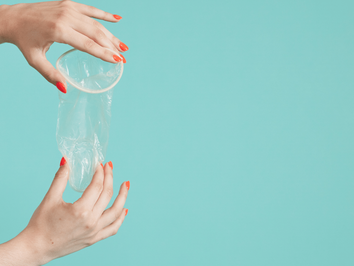 A woman with red nail polish holding a female condom with both hands against a plain turquoise background. One hand is gripping the top rim, while the other supports the bottom of the condom.